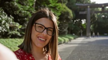 Gözlüklü neşeli genç İspanyol kadın bu güzel resimde aşk ve mutluluk saçan Japonya 'nın ikonik meiji tapınağındaki selfie ile kalp şeklindeki el işaretini yakalıyor.