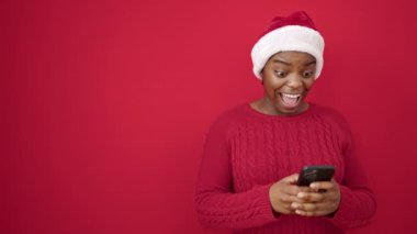 Afrika kökenli Amerikalı bir kadın Noel şapkası takıyor ve izole edilmiş kırmızı arka plan üzerinden akıllı telefon kullanıyor.