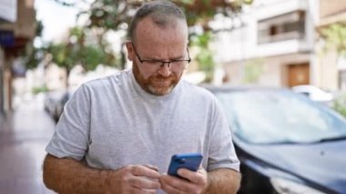 Neşeli, gözlüklü, sakallı, orta yaşlı, beyaz bir adam canlı, şehir sokağında telefonundan bangır bangır müzik dinliyor
