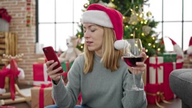 Genç sarışın kadın akıllı telefon kullanıyor evde Noel 'i kutlamak için şarap içiyor.