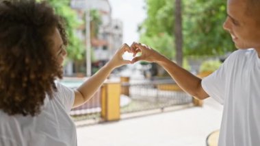 Güzel bir açık hava parkında, bir çift yan yana durur, elleri kalp şeklinde bir jest yapar. Mutlu, kendinden emin gülümsemeleri güneşli gökyüzünün altında neşe saçıyor..