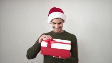 Genç, İspanyol bir adam hediyesini açıyor, beyaz arka planda noel şapkası takıyor.