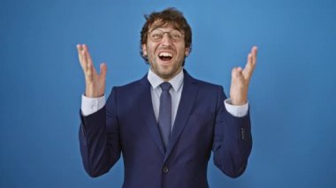 Heyecanlı takım elbiseli genç adam çıldırıyor, zaferi kollarıyla kutluyor. İzole edilmiş mavi arka planda neşeyle köpürüyor. Zafer hiç bu kadar yakışıklı olmamıştı..