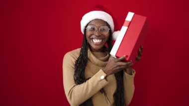 Afrikalı bir kadın, kırmızı arka planda kendine güvenen bir şekilde Noel hediyesini tutuyor.