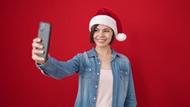 Kafkasyalı genç bir kadın izole edilmiş kırmızı arka planda noel şapkası takarak selfie çekiyor.