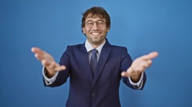 Mavi takım elbiseli ve gözlüklü yakışıklı genç sakallı adam dostça yardım eli uzatıyor, olumlu bir hava yayıyor. İzole edilmiş mavi arka plan üzerinde davetkar bir jest. Gel ve başarıya katıl.!