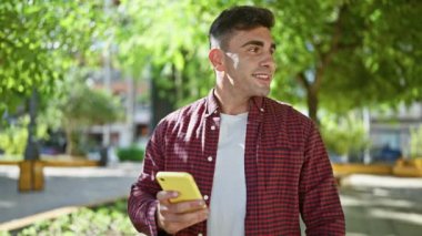 Neşeli genç İspanyol adam parkta doğanın tadını çıkarıyor, akıllı telefonu tutuyor ve kullanıyor, güzel bir gülümsemeyle mutlu bir şekilde kısa mesaj yazıyor.