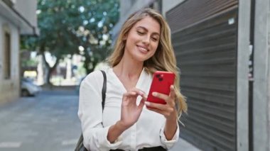 Neşeli ve çekici sarışın kadın zaferini dışarıda kutluyor. Canlı bir şehir caddesinde telefonunu neşeyle kullanıyor. Kazandığını mesaj atarken gülümsüyor..