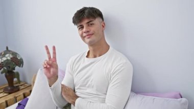 Yatak odası fotoğrafı, gülümseyen İspanyol genç adam pijama giyerek yatakta yatıyor. Mutlu yüz göz kırpıyor ve iki parmaklı zafer işareti parlıyor.