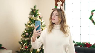 Genç sarışın kadın akıllı telefonuyla selfie çekiyor evde Noel ağacının yanında duruyor.