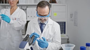 Bilimden iki gülümseyen bilim adamı, kapalı bir laboratuardaki pipetle test tüpüne sıvı dökerek, yeni tıbbi analizleri keşfederken güvenlik eldivenlerini yakınlaştırırlar..