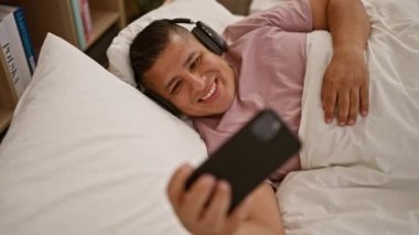 Neşeli genç latin adam neşeli bir şekilde el sallıyor. Mobil bir video araması sırasında, yatak odasında uzanıyordu. Arka planda görünen iletişim teknolojisini kullanıyordu.