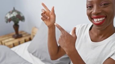 Neşeli Afro-Amerikalı kadın rahat bir yatakta uzanıyor, kendinden emin bir şekilde yan tarafı gösteriyor, güzel bir yatak odasındaki kameraya hoş bir gülümsemeyle bakıyor..