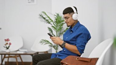 Genç latin adam, telefonuna dalmış, kulaklıklarından ritmi dinliyor, bekleme odasındaki sandalyeye oturmuş.