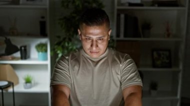 Gülümseyen genç latin adam ofiste rahatça dinleniyor, elleri başının üstünde dinleniyor, geç saatlere kadar dizüstü bilgisayarla çalışırken.