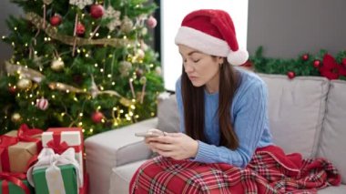 Kafkasyalı genç bir kadın Noel 'i evde üzgün bir ifadeyle akıllı telefon kullanarak kutluyor.