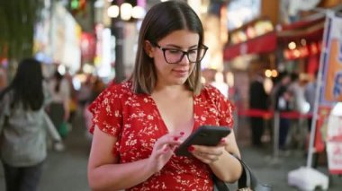 Gözlüklü, neşeli, güzel İspanyol kadın Osaka 'nın ünlü Dotonbori Caddesi' nde akıllı telefonuyla gece mesajlaşmasının tadını çıkarıyor.