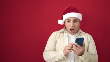 Genç İspanyol adam yılbaşı şapkası takıyor ve izole edilmiş kırmızı arka plan üzerinde akıllı telefon kullanıyor.