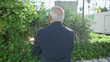 Neşeli, gri saçlı, neşeli bir şekilde şehir parkında gülümseyen, şık bir takım elbise ve güneş gözlüğü takan, pozitif duruşuyla duran, kolları güvenle çaprazlanmış, neşeli bir son sınıf öğrencisi..
