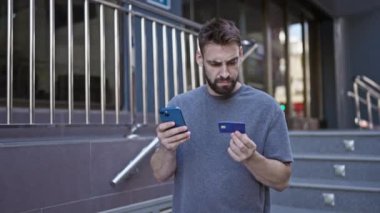 Akıllı telefonuna bakan, şehir caddesinde kredi kartı kullanarak online alışveriş ödemesiyle boğuşan genç İspanyol bir adam.