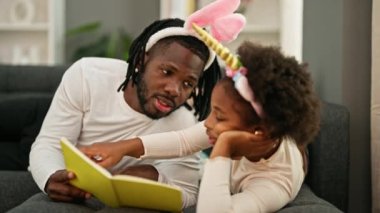 Afrika kökenli Amerikalı baba ve kız evdeki kanepede uzanmış kitap okuyorlar.