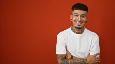 Kendinden emin, dövmeli, kolları çapraz pozlar veren, kameraya sevinçle gülümseyen, kırmızı duvarda izole edilmiş genç bir adam.