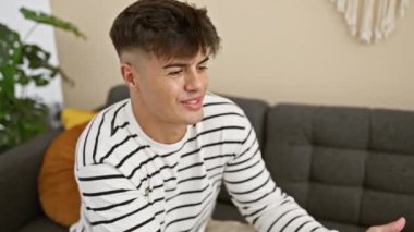 Gülümseyen İspanyol genç adam evdeki bir kanepede rahatça otururken eğlenceli bir sohbetten zevk alıyor, neşe, güven ve çekici, rahatlatıcı bir aura yayıyor.