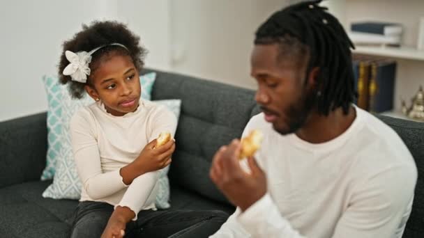 アフリカ系アメリカ人の父親と娘がソファーに座って自宅でクロワッサンを食べる — ストック動画