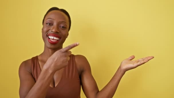 アフリカ系アメリカ人女性は驚きに立ち 明るく微笑み 孤立した黄色の背景にある空間を指差しながら 自信を持って手で提示します — ストック動画