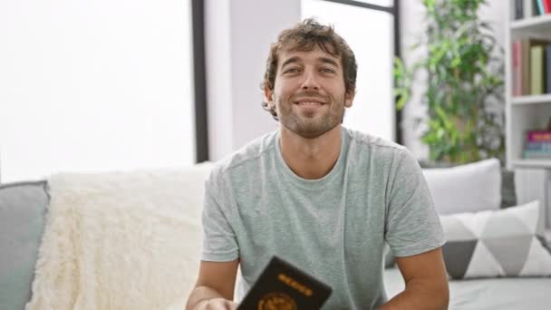 喜んで若いヒスパニックの男は自信を持って微笑み メキシコのパスポートを持って 自宅のリビングルームのソファーに座って 彼の休暇旅行を計画することに興奮 — ストック動画