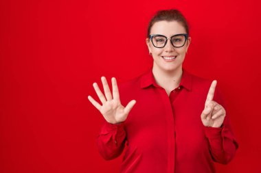 Kırmızı arka planda kızıl saçlı genç İspanyol kadın altı numaralı parmaklarıyla gülümseyerek mutlu ve kendinden emin bir şekilde duruyor.. 