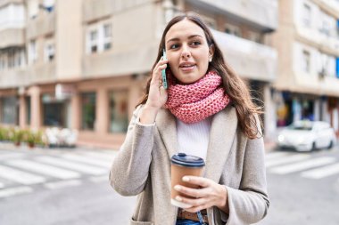 Young beautiful hispanic woman talking on smartphone drinking coffee at street