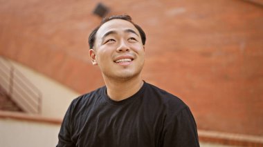 Neşeli genç Çinli adam, kendinden emin bir şekilde gülümsüyor, şehir caddesinde dururken gökyüzüne bakıyor.