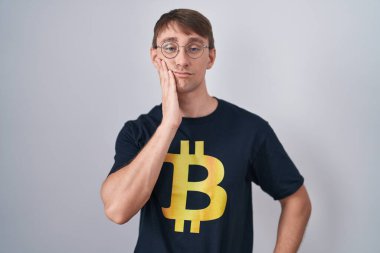 Bitcoin tişört giyen beyaz tenli sarışın adam kollarını kavuşturarak depresyondan sıkılmış ve yorgun görünüyor.. 