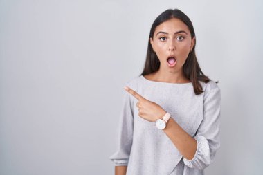 Young hispanic woman standing over white background surprised pointing with finger to the side, open mouth amazed expression. 
