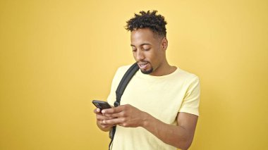 Sırt çantası takan Afrikalı Amerikalı bir adam izole edilmiş sarı arka plan üzerinde akıllı telefon kullanıyor.