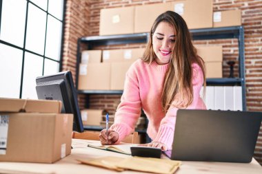 Young beautiful hispanic woman ecommerce business worker using laptop and calculator writing on notebook at office