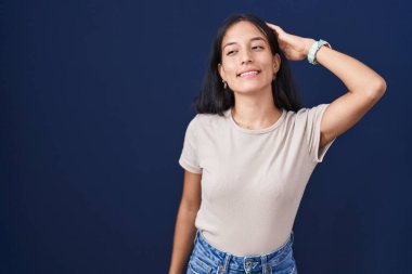 Young hispanic woman standing over blue background smiling confident touching hair with hand up gesture, posing attractive and fashionable 