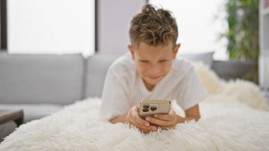 Sevimli sarışın çocuk, neşe dolu, akıllı telefonunu neşeyle kullanıyor, evde, kanepede yatarken mesajlaşıyor.