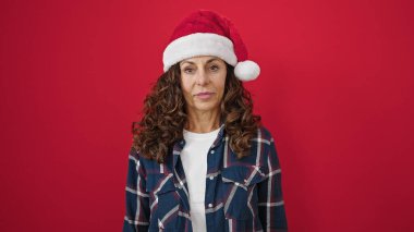 Orta yaşlı İspanyol kadın, izole edilmiş kırmızı arka planda ciddi bir ifadeyle noel şapkası takıyor.