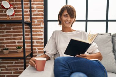Young beautiful hispanic woman reading book and drinking coffee sitting on sofa at home