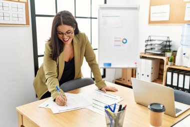 Young beautiful hispanic woman business worker writing on document standing at office