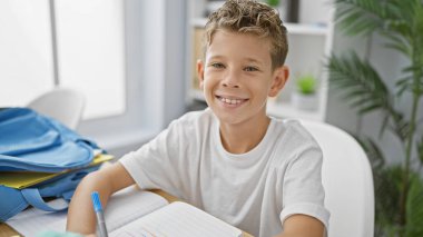 Masasında notlar almaya dalmış neşeli sarışın çocuk. Sınıfın atmosferinde gülümsüyor, akademik coşku sergiliyor..