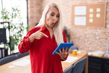 Ofiste çalışan beyaz bir kadın. Tablet parmaklarını göstererek reklamını yapıyor. Şaşırmış bir yüz ve açık bir ağızla. 