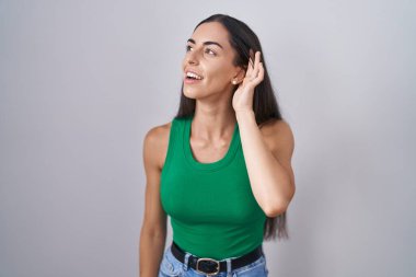 Young woman standing over isolated background smiling with hand over ear listening an hearing to rumor or gossip. deafness concept. 