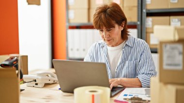 Orta yaşlı bir kadın, ofis masasında dizüstü bilgisayar kullanarak iş yapıyor.