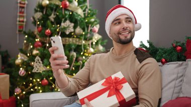 Genç Arap adam elinde akıllı telefon hediyesiyle evde Noel ağacının yanında oturuyor.