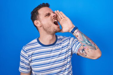 Young hispanic man standing over blue background shouting and screaming loud to side with hand on mouth. communication concept. 
