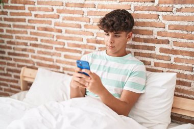 Latin kökenli genç, yatak odasında rahat bir ifadeyle akıllı telefon kullanıyor.