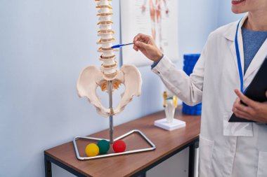 Genç sarışın kadın pisiyoterapist rehabilitasyon kliniğinde elinde pano tutan omurların anatomik modelini gösteriyor.